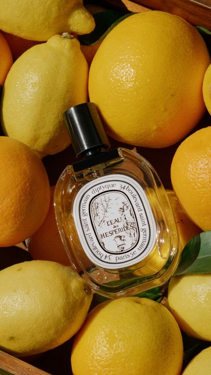 A bottle of Diptyque L'Eau des Hespérides resting on a crate of lemons and oranges