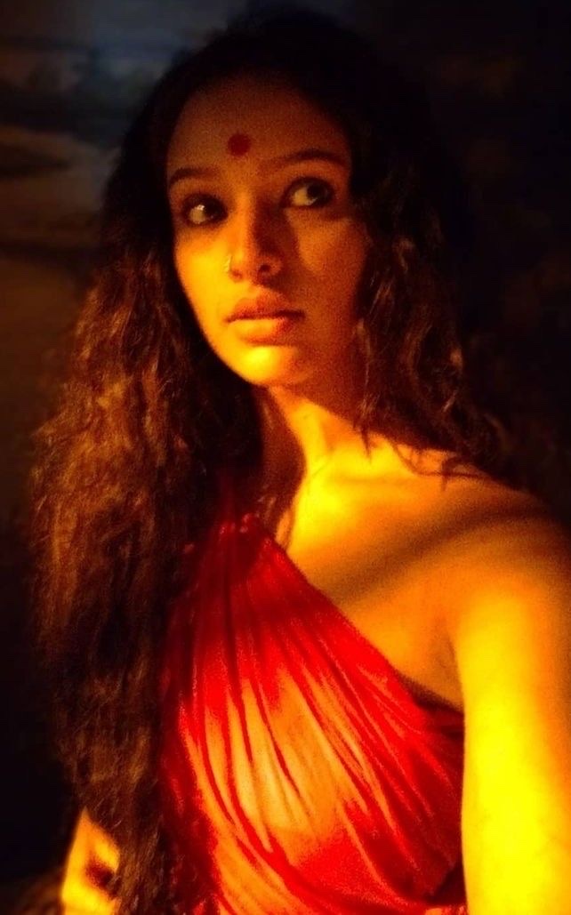 A woman with a red bindi and loose curls, draped in vermilion cloth and lit by warm firelight