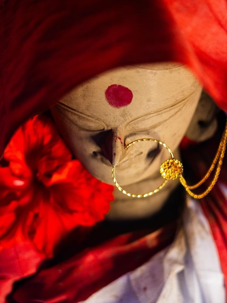 A clay devi idol with a red bindi and gold nose ring, framed by red hibiscus and cloth